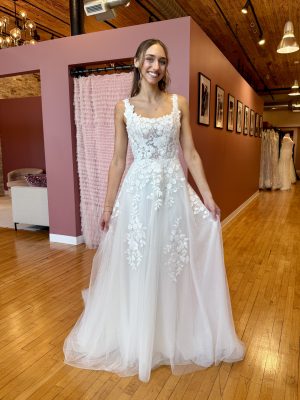 Brunette woman in a white lace wedding gown in a boutique with pink walls and framed photos.