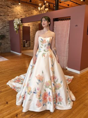 Young woman in strapless floral ball gown in boutique with brick walls and pink decor.